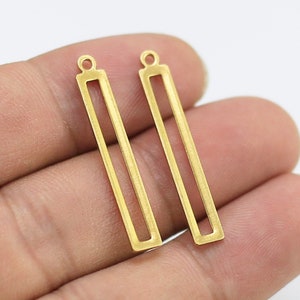 Peut inclure: Deux pendentifs rectangulaires en métal de couleur dorée avec une boucle en haut. Les pendentifs sont ouverts sur les côtés et ont une finition lisse.