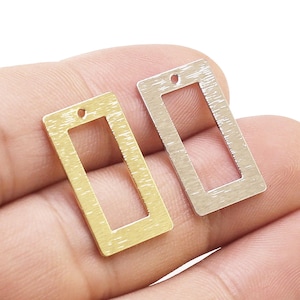 May include: Two rectangular metal charms with a brushed finish, one in a gold tone and one in a silver tone. Each charm features a hollow centre and a small circular hole at the top for attachment, presenting a flat, geometric design.