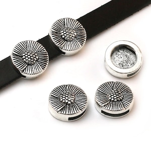 May include: Silver-coloured metal sunflower-shaped jewellery findings. The findings are round with a detailed floral design and a small loop on the back. A black leather strap is visible, with two findings attached. The findings are scattered on a white surface.