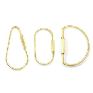 Könnte beinhalten: Drei goldene Schlüsselanhänger in verschiedenen Formen: eine Träne, ein Oval und ein D. Jeder Schlüsselanhänger hat einen sechseckigen Ring.
