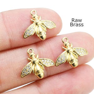 May include: Three gold-colored bee charms with crystal-like accents on the wings. The charms are small and detailed, with a loop at the top for attaching to jewelry. The text "Raw Brass" is visible.
