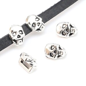 May include: Silver skull charm beads for jewelry making. The beads are detailed with skull faces and are shown on a black leather cord.