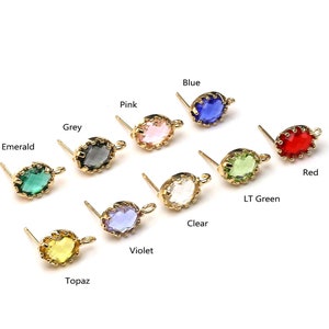 Puede incluir: Una colección de pendientes de botón dorados con piedras preciosas ovaladas de colores. Las piedras son de varios colores, incluyendo verde esmeralda, gris, rosa, azul, rojo, verde claro, transparente, violeta y topacio. Cada pendiente tiene un pequeño lazo.