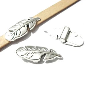 May include: Three silver-toned metal feather-shaped sliders for leather cord. One slider is attached to a flat tan leather strip, while two additional loose feather sliders are shown, one displaying the flat back and one showing the front.