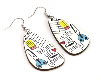 May include: Teardrop-shaped earrings with a teacher theme. Each earring features a white background with a pencil, scissors, coffee cup, and the words "Teacher Life Coffee." The earrings have silver-colored hooks.