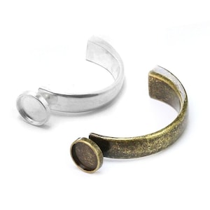 Könnte beinhalten: Zwei Ringrohlinge aus Silber und Bronze mit einer runden Fassung zum Einsetzen eines Cabochonsteins. Die Ringrohlinge sind an einer Seite offen und haben eine glatte Oberfläche.