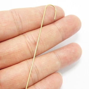 May include: A single, slender, gold-colored ear cuff earring. The earring has a straight, thin post that curves at the top to form a hook. The earring is shown against a white background.