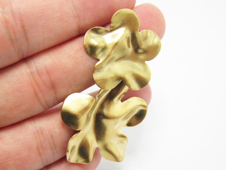 K&ouml;nnte beinhalten: Zwei goldfarbene Metall-Blumenanh&auml;nger mit strukturierter Oberfl&auml;che. Die Anh&auml;nger sind an der Basis verbunden und haben eine leicht unregelm&auml;&szlig;ige Form.