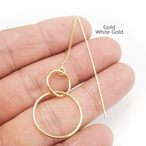 Puede incluir: Un pendiente de hilo dorado con dos aros circulares entrelazados suspendidos de una cadena fina. El diseño incluye un poste de metal recto para su inserción. El texto Gold White Gold es visible en la esquina superior derecha de la imagen.