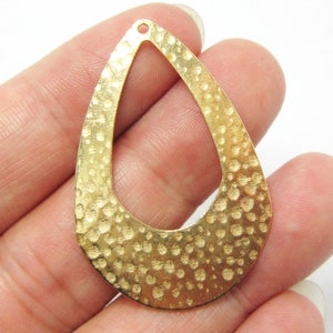 May include: A gold-colored, hammered-texture teardrop-shaped earring component. The earring has a small hole at the top for attachment. The surface has a dimpled texture, giving it a rustic appearance. The earring is held by a hand.