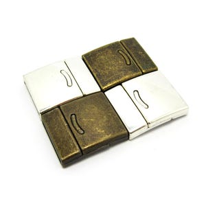 Puede incluir: Cuatro cierres magnéticos cuadrados dispuestos en un patrón de cuadrícula. Dos cierres tienen un acabado plateado pulido y dos cierres tienen un acabado de bronce antiguo. Cada cierre presenta una superficie rectangular plana con un detalle.