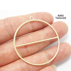 May include: A gold-toned metal pendant featuring a circular frame with a horizontal bar across the center and a small loop at the top for attachment. The surface has a fine textured finish and the text R495 Textured is printed in black.