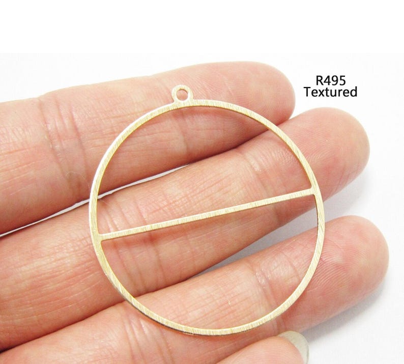 May include: A gold-toned metal pendant featuring a circular frame with a horizontal bar across the center and a small loop at the top for attachment. The surface has a fine textured finish and the text R495 Textured is printed in black.