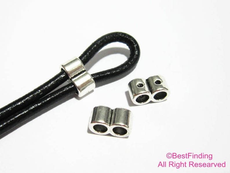 May include: Two silver metal cord end stoppers for leather cords. The stoppers are shaped like a double circle. A black leather cord is threaded through one of the stoppers.