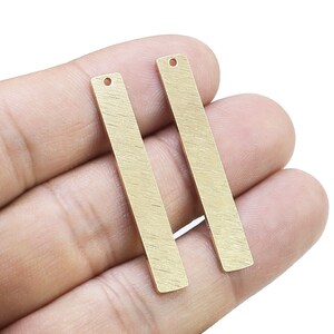 Puede incluir: Dos colgantes de barra de metal de color dorado con un agujero en la parte superior. Las barras están cepilladas y tienen una superficie ligeramente texturizada.