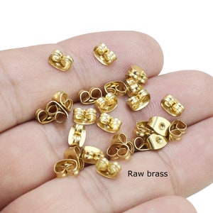 Puede incluir: Primer plano de un montón de respaldos de pendientes de color dorado. Los respaldos son de metal y tienen un diseño sencillo. El texto "Raw brass" es visible en la imagen.