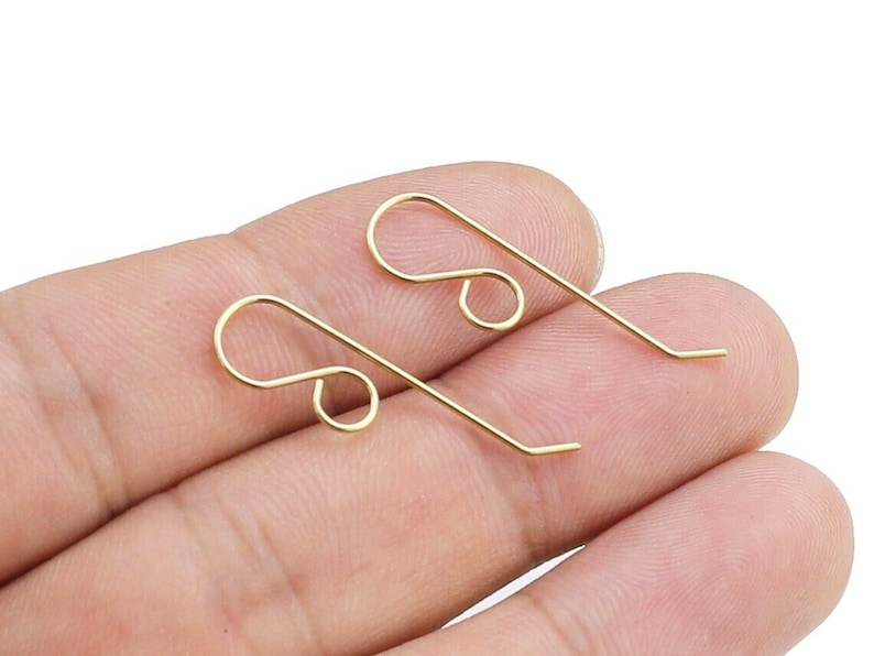 May include: Two gold-toned earring hooks, also known as ear wires, are shown on a hand. The hooks are made of metal and have a simple, classic design.