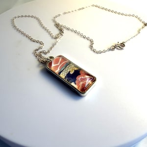 May include: A silver necklace with a rectangular pendant. The pendant features a colorful floral design with red, blue, and gold hues. The chain is delicate and the clasp is visible.