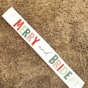 May include: White satin sash with the text "Merry and Bride" in colorful, stylized letters. The letters are in shades of red, pink, green, and white.