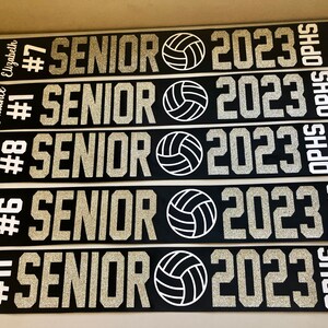 Puede incluir: Cinco fajas negras y blancas con texto de purpurina plateada. Cada faja dice "Senior 2023" con un gr&aacute;fico de voleibol y un n&uacute;mero y un nombre. Las fajas son para "Hannah #11", "Izzy #6", "Dylan #8", "Cadence #1" y "Elizabeth #7".