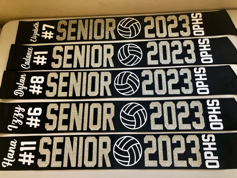 Puede incluir: Cinco fajas negras y blancas con texto de purpurina plateada. Cada faja dice "Senior 2023" con un gr&aacute;fico de voleibol y un n&uacute;mero y un nombre. Las fajas son para "Hannah #11", "Izzy #6", "Dylan #8", "Cadence #1" y "Elizabeth #7".