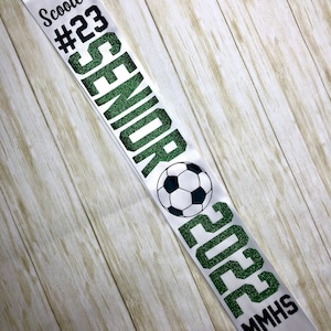 Puede incluir: Una banda de graduaci&oacute;n blanca con texto verde brillante que dice "Scooter #23 Senior 2023 MMHS" y un gr&aacute;fico de bal&oacute;n de f&uacute;tbol.