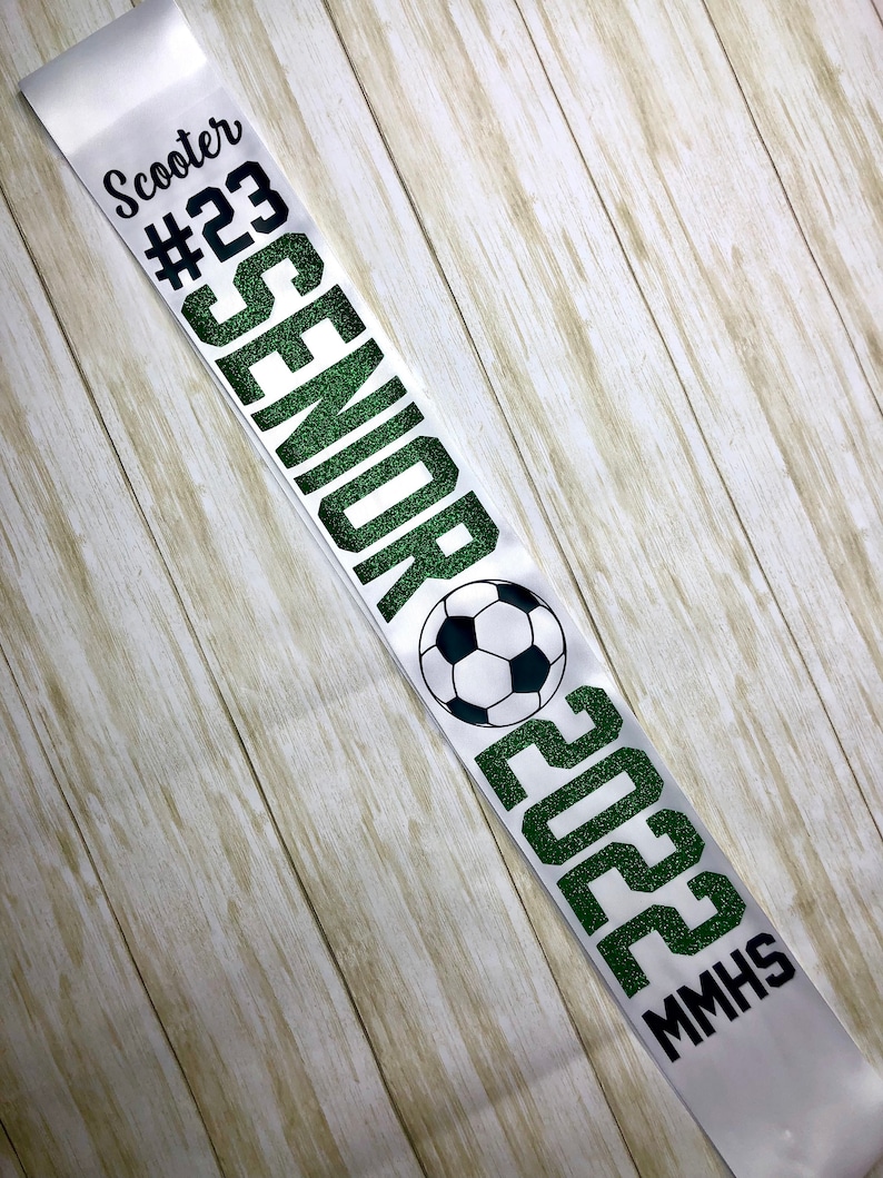 Puede incluir: Una banda de graduaci&oacute;n blanca con texto verde brillante que dice "Scooter #23 Senior 2023 MMHS" y un gr&aacute;fico de bal&oacute;n de f&uacute;tbol.