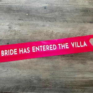 May include: Pink satin sash with the text "THE BRIDE HAS ENTERED THE VILLA" in white lettering and a gold glitter heart.