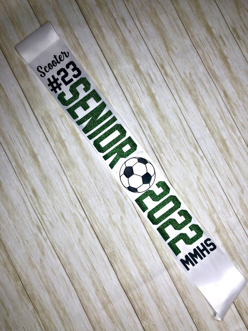 Puede incluir: Banda de sat&eacute;n blanco con texto verde brillante que dice "Senior 2023 MMHS" y un gr&aacute;fico de bal&oacute;n de f&uacute;tbol. La banda tambi&eacute;n incluye el texto "Scooter #23".