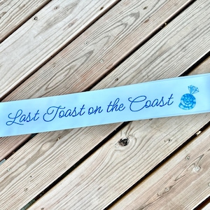 Könnte beinhalten: Ein hellblaues Satinsband mit dem Text "Last Toast on the Coast" in blauer Kursivschrift. Auf der rechten Seite des Bandes befindet sich eine stilisierte Illustration eines Rings in Blau und Weiß.