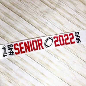 Puede incluir: Pancarta blanca y roja con el texto "Senior 2022" y un gr&aacute;fico de f&uacute;tbol. La pancarta tambi&eacute;n incluye el texto "Nicole #49 SRHS".