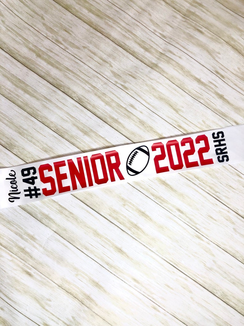 Puede incluir: Pancarta blanca y roja con el texto "Senior 2022" y un gr&aacute;fico de f&uacute;tbol. La pancarta tambi&eacute;n incluye el texto "Nicole #49 SRHS".
