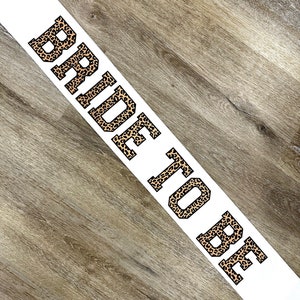 Peut inclure: Sash en satin blanc avec l'inscription "Bride to Be" imprimée en lettres noires avec un motif léopard.