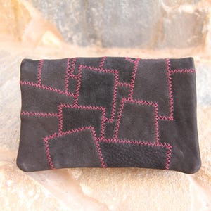 Könnte beinhalten: Eine schwarze Leder-Clutch mit Patchwork-Design. Die Clutch ist mit pinkfarbenem Garn zusammengenäht.