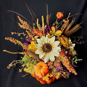 May include: A fall-themed centerpiece featuring a faux pumpkin, sunflowers, and various dried flowers and greenery. The arrangement is in shades of orange, yellow, brown, and purple.