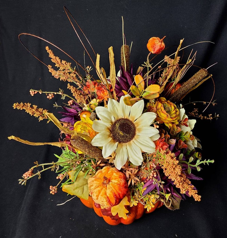 May include: A fall-themed centerpiece featuring a faux pumpkin, sunflowers, and various dried flowers and greenery. The arrangement is in shades of orange, yellow, brown, and purple.