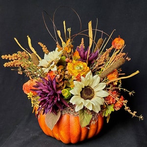 May include: A fall-themed arrangement featuring a faux pumpkin filled with a variety of dried flowers and greenery. The arrangement includes sunflowers, purple flowers, and orange and yellow flowers.