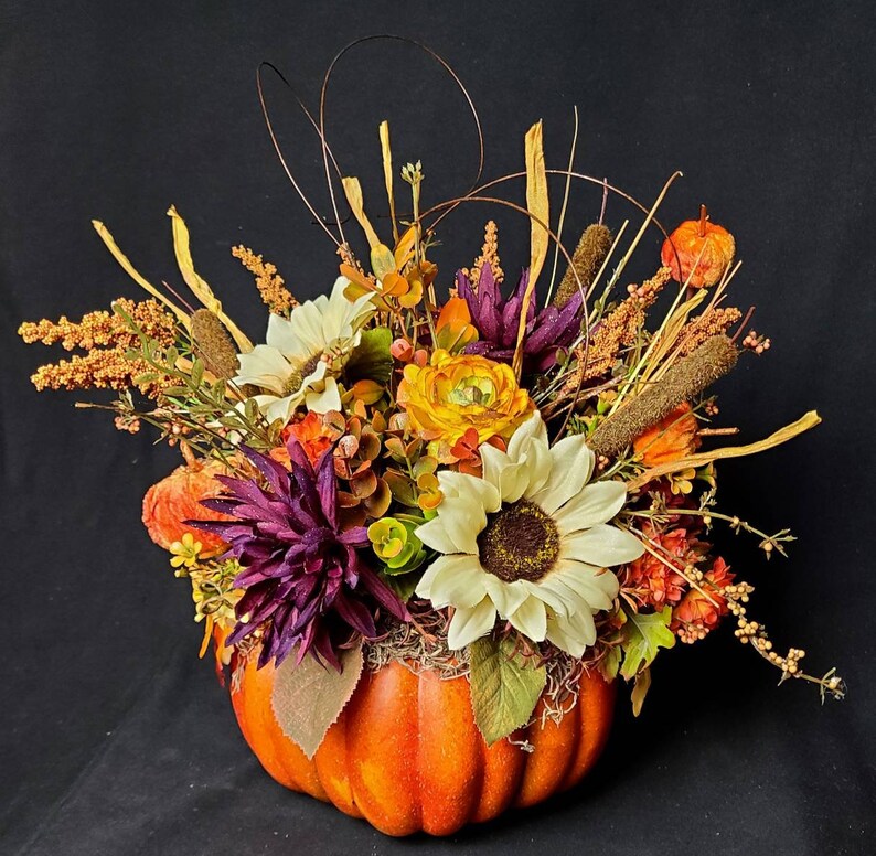 May include: A fall-themed arrangement featuring a faux pumpkin filled with a variety of dried flowers and greenery. The arrangement includes sunflowers, purple flowers, and orange and yellow flowers.