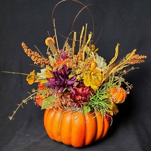 May include: A fall-themed floral arrangement in a pumpkin. The arrangement features a variety of flowers in shades of yellow, orange, purple, and white, as well as dried grasses and twigs.