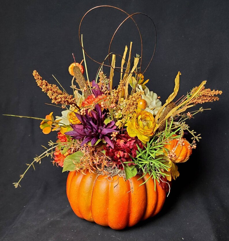 May include: A fall-themed floral arrangement in a pumpkin. The arrangement features a variety of flowers in shades of yellow, orange, purple, and white, as well as dried grasses and twigs.