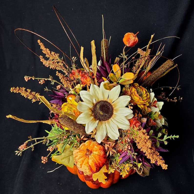 May include: A fall-themed floral arrangement in a pumpkin. The arrangement features a large yellow sunflower, smaller yellow flowers, purple and green foliage, and brown twigs.