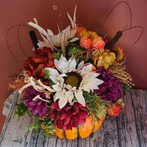 May include: A fall-themed centerpiece featuring a pumpkin filled with a variety of artificial flowers, including white sunflowers, burgundy chrysanthemums, and purple flowers. The arrangement also includes dried grasses and wheat.