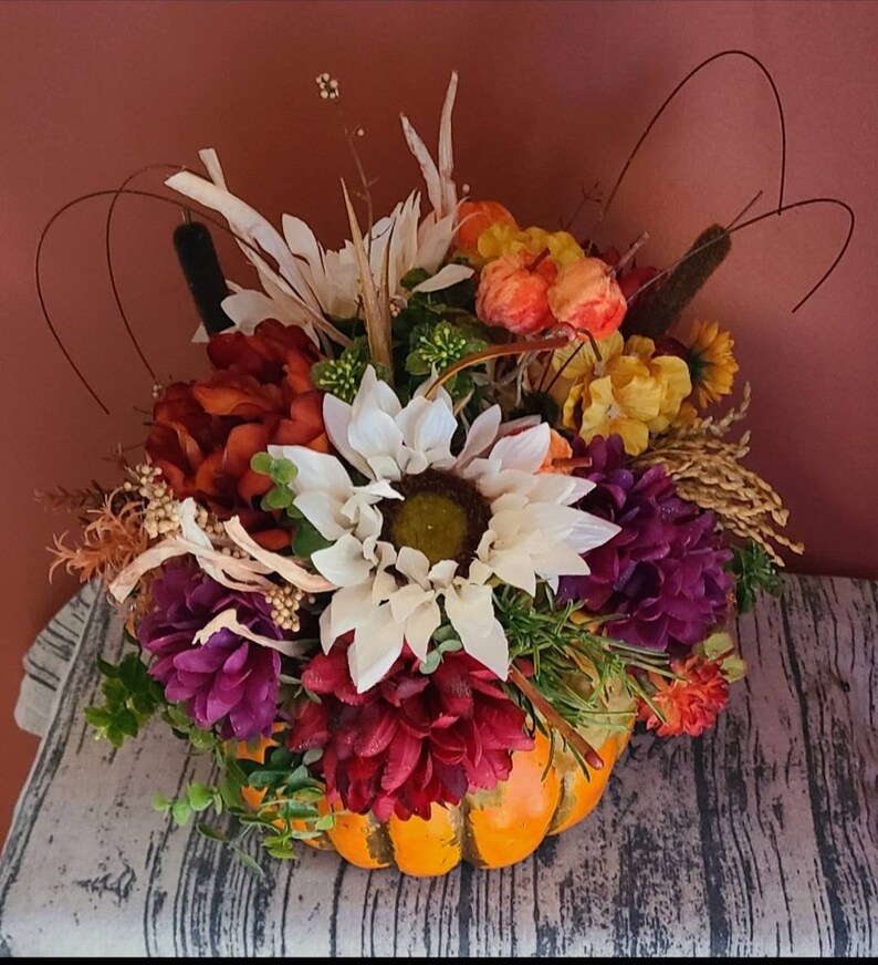 May include: A fall-themed centerpiece featuring a pumpkin filled with a variety of artificial flowers, including white sunflowers, burgundy chrysanthemums, and purple flowers. The arrangement also includes dried grasses and wheat.