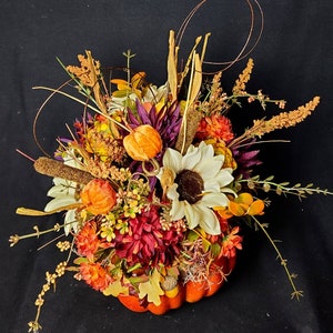 May include: A fall-themed centerpiece featuring a pumpkin filled with a variety of flowers, including sunflowers, mums, and berries. The arrangement is accented with dried grasses and twigs.