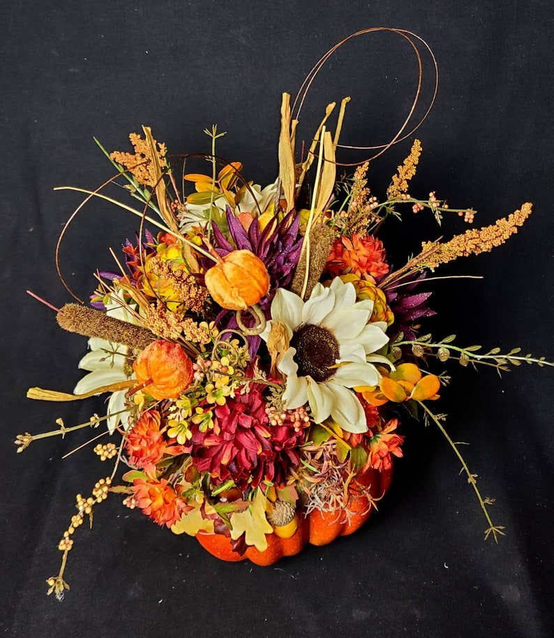 May include: A fall-themed centerpiece featuring a pumpkin filled with a variety of flowers, including sunflowers, mums, and berries. The arrangement is accented with dried grasses and twigs.