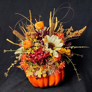 May include: A fall-themed floral arrangement in a bright orange pumpkin. The arrangement features a variety of flowers, including sunflowers, mums, and berries, in shades of orange, yellow, red, and white.