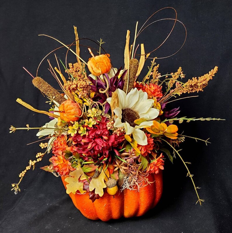 May include: A fall-themed floral arrangement in a bright orange pumpkin. The arrangement features a variety of flowers, including sunflowers, mums, and berries, in shades of orange, yellow, red, and white.