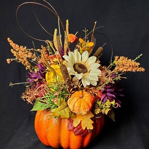 May include: A fall-themed arrangement featuring a faux pumpkin filled with dried flowers, leaves, and berries. The arrangement includes a white sunflower, orange berries, and brown dried grasses.