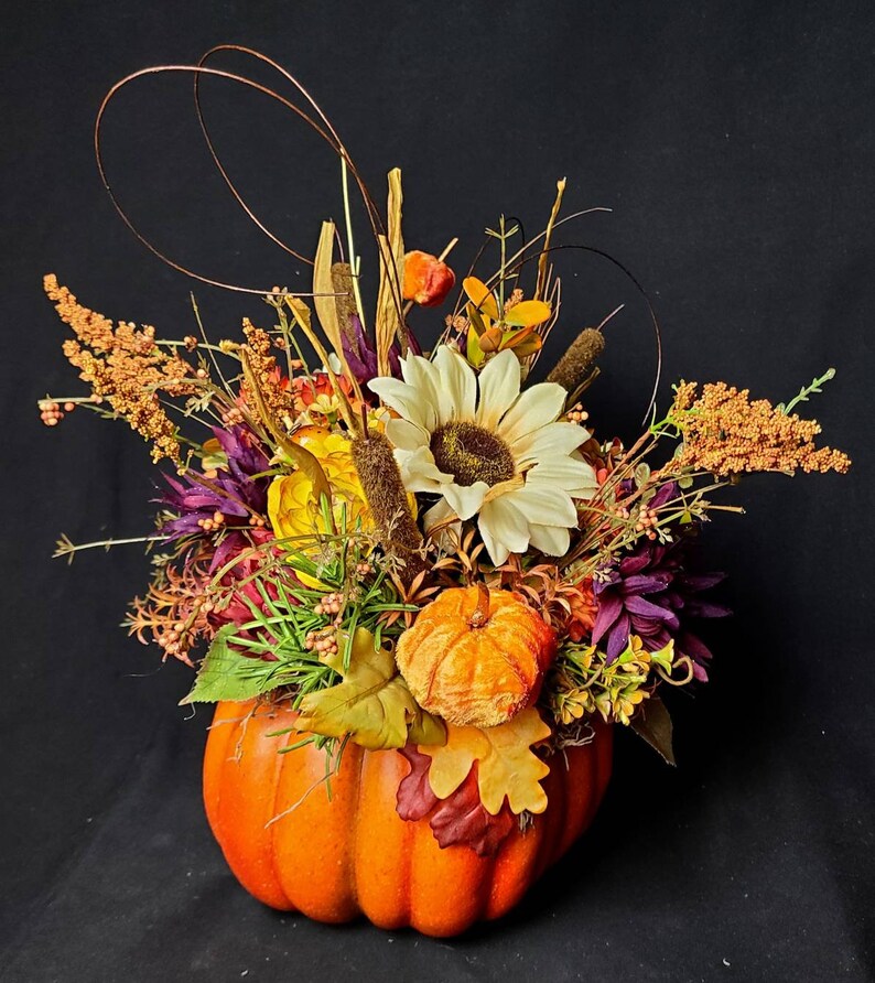 May include: A fall-themed arrangement featuring a faux pumpkin filled with dried flowers, leaves, and berries. The arrangement includes a white sunflower, orange berries, and brown dried grasses.