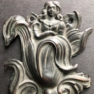May include: A bronze decorative wall plaque featuring a woman in a flowing gown, surrounded by intricate floral designs.